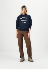 Tengerészkék pulóver, rajta fehér hímzéssel a "Timberland Authentic Est 1973" felirattal, barna nadrággal és szürke, fekete fűzős csizmával párosítva.