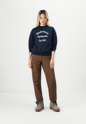 Marineblauer Pullover mit "Timberland Authentic Est 1973" in Weiß gestickt, kombiniert mit braunen Hosen und grauen Stiefeln mit schwarzen Schnürsenkeln.