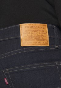 Pantalones vaqueros de mezclilla azul oscuro con un parche de cuero marrón claro. El parche presenta texto e imágenes en relieve, incluyendo caballos y el nombre de la marca.