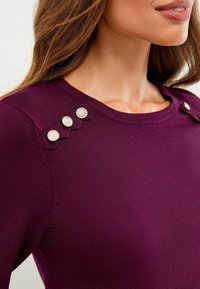 Haut tricot violet avec un col rond et quatre boutons métalliques décoratifs sur l'épaule gauche, en tissu lisse et texturé.