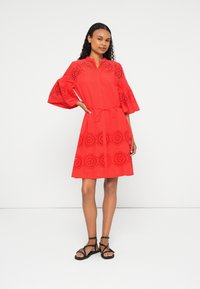 Vestido vermelho com mangas de sino, padrões recortados redondos nas mangas e na bainha, cinto ajustável na cintura e tecido leve. Usado com sandálias pretas.