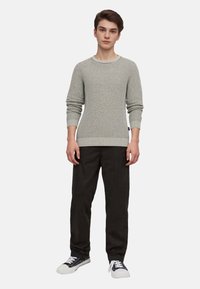 Hellgrauer strukturierter Strickpullover mit rundem Hals und gerippten Bündchen, kombiniert mit schwarzen Hosen und schwarz-weißen Sneakers.