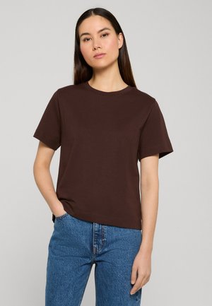 Jeune femme aux longs cheveux bruns portant un t-shirt marron foncé uni à manches courtes et un jean bleu, main dans la poche, fond neutre.