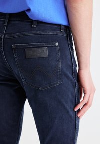 Temno modri traper hlače s usnjenim Wrangler logotipom na zadnjem žepu, s kontrastnim šivi in klasičnim dizajnom s petimi žepi.