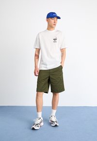Hvid t-shirt med et grafisk logo, olivengrønne shorts, blå kasket og hvide sneakers med sorte og orange accenter. Glat stof og afslappet pasform.