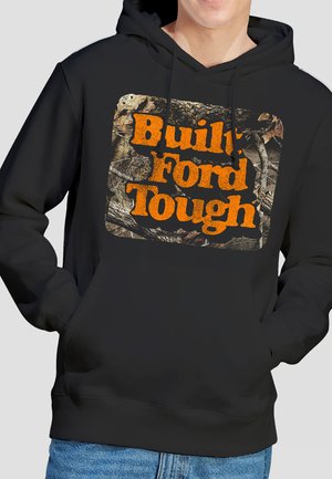 Schwarzer Kapuzenpullover mit einem Tarnmuster-Quadrat und orangem Text "Built Ford Tough", getragen von einer Person mit den Händen in den vorderen Taschen, dazu sichtbare blaue Jeans.
