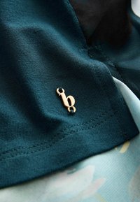 Teflonnene Baumwollstoffe in Türkis mit glatter Textur sind mit einem kleinen goldenen Logo akzentuiert. Der Hintergrund zeigt ein weiches, hell gemustertes Material.