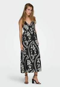 Maxi-jurk van zwarte stof met een groot wit bloemenpatroon, V-halslijn en een aangesloten taille, gecombineerd met zwarte sandalen met bandjes.