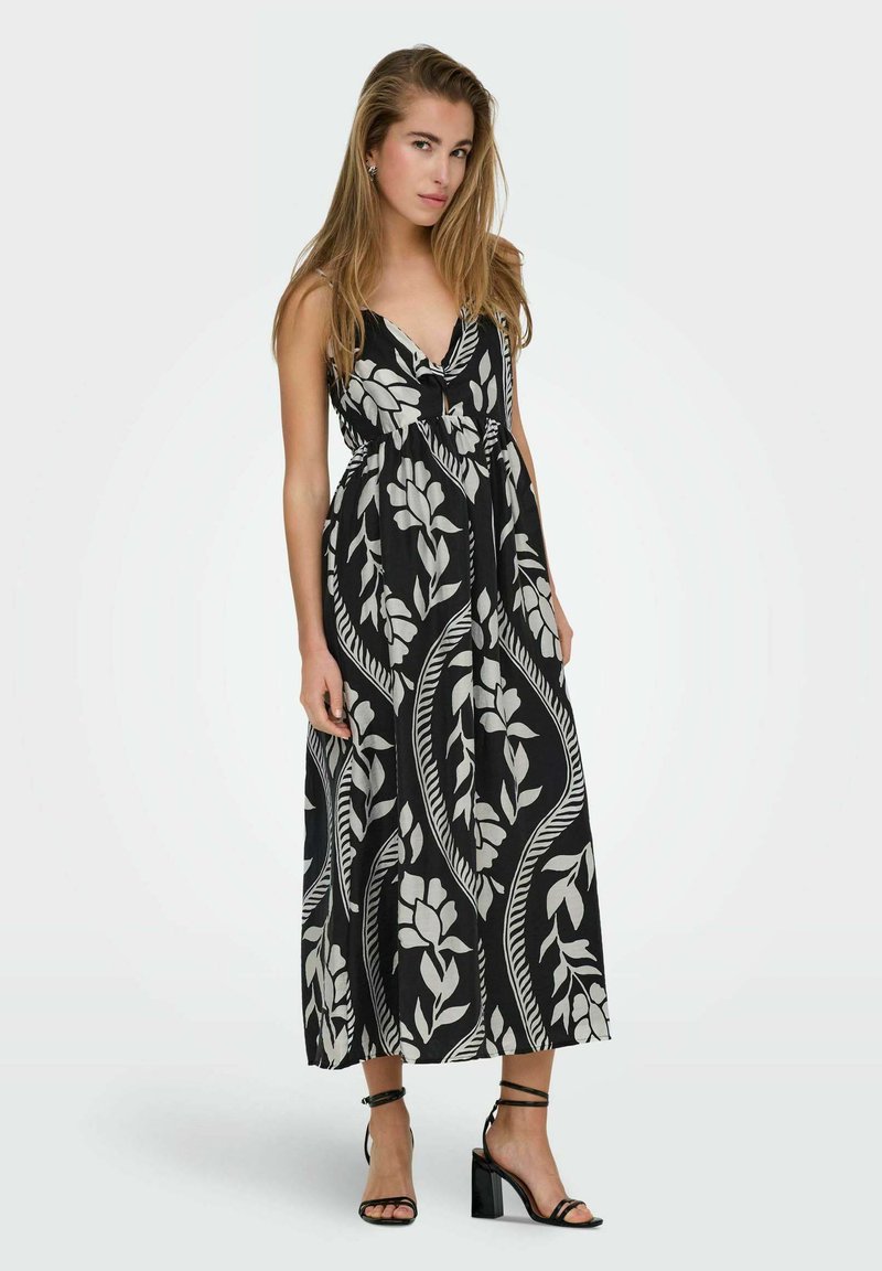 Maxi-jurk van zwarte stof met een groot wit bloemenpatroon, V-halslijn en een aangesloten taille, gecombineerd met zwarte sandalen met bandjes.