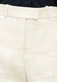 Κοντινό πλάνο από κρεμ παντελόνι ψηλόμεσο με ορατές ραφές και θηλιές για ζώνη, σε συνδυασμό με σκούρο ρούχο με υφή.