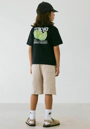 Kind draagt een zwarte pet, zwart T-shirt met limegroene afbeelding en "Tokyo" tekst, beige korte broek, witte sokken en beige sandalen, staand zijwaarts.
