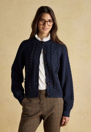 Młoda kobieta z długimi ciemnymi włosami, nosząca duże okulary, granatowy sweter w warkocze, białą bluzkę i brązowe spodnie w kratę, stoi z rękami w kieszeniach.