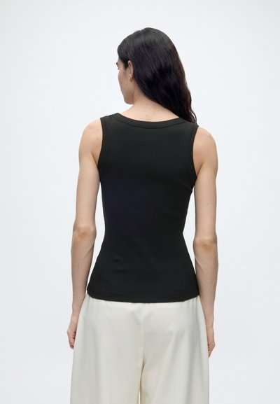 Femme aux longs cheveux foncés portant un haut noir sans manches et un pantalon blanc ample, debout de dos face à la caméra sur un fond uni.