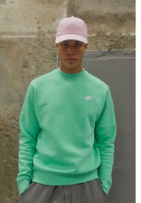 Sweatshirt verde menta com punhos e gola canelados, apresentando um pequeno logo branco da Nike. Acompanhado por um boné de beisebol rosa claro.