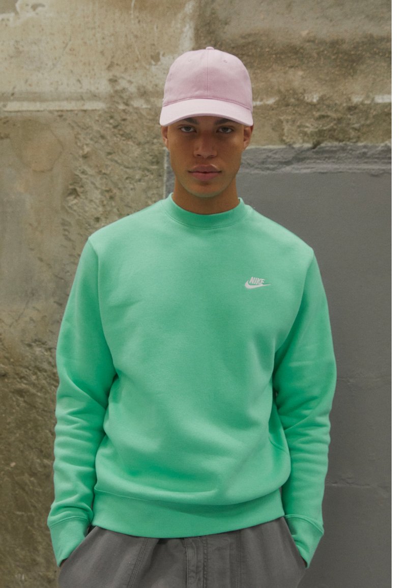 Sweatshirt verde menta com punhos e gola canelados, apresentando um pequeno logo branco da Nike. Acompanhado por um boné de beisebol rosa claro.