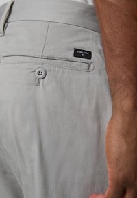 Ljust grå byxor i mjuk kvalitet har en bakficka med knapp och en svart loggohängare sytt på midjebandet.