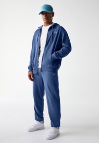 Haut à zip bleu avec logo LA, associé à un pantalon de survêtement bleu assorti. Complété par une casquette bleu clair et des baskets blanches. Texture lisse.