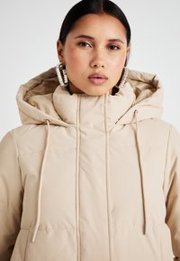 Doudoune beige avec un col montant et une capuche, dotée de cordons de serrage. Surface lisse avec des sections matelassées, accompagnée de grandes boucles d'oreilles en forme de cercle.