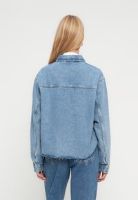 Blond person iført lys blå denimjakke og jeans, stående med ryggen mot kameraet foran en hvit bakgrunn.