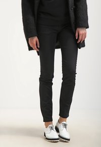 Svarta tajta byxor i kombination med vita skor med svarta detaljer och snören. Outfitten inkluderar en texturerad svart kappa.