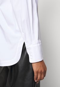 Camicia bianca a maniche lunghe con polsini abbottonati, caratterizzata da una texture liscia e slittini laterali, abbinata a pantaloni in pelle nera.
