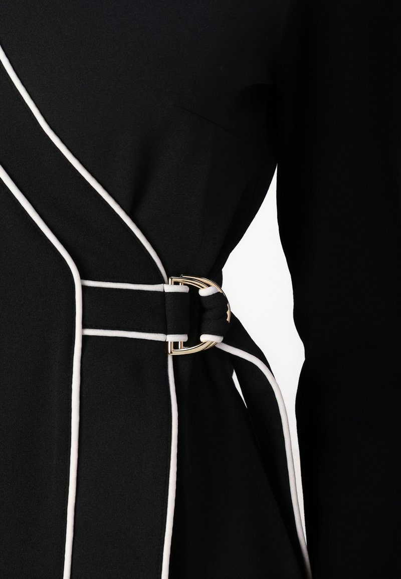 Vestido negro con ribetes blancos, que presenta un cinturón con hebilla en tono dorado en la cintura. La tela tiene una textura suave y un corte ajustado.