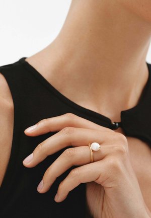 Mano con piel suave que lleva un delicado anillo de oro con una perla blanca, descansando cerca del cuello con una blusa negra sin mangas.