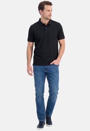 Man die een zwart poloshirt met korte mouwen, een blauwe spijkerbroek en donkere sneakers draagt, staat met één hand in zijn zak en kijkt naar rechts.