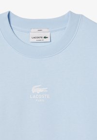 Svjetloplava uniseks dukserica s okruglim izrezom, koja ima teksturiranu tkaninu i bijeli Lacoste logo te natpis otisnut u sredini.