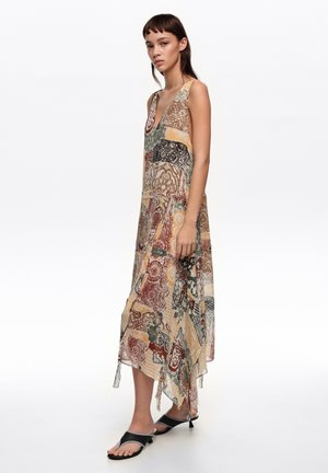 Mujer con vestido sin mangas de patrón patchwork y bajo asimétrico, calzando sandalias negras de tacón bajo, posando frente a un fondo blanco.