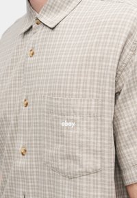 Πουκάμισο με κοντά μανίκια, ανοιχτό καφέ καρό, με γιακά που κουμπώνει, ξύλινα κουμπιά και τσέπη στο αριστερό στήθος με λευκό λογότυπο.