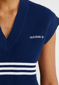 Ναυτικό ριγέ πλεκτό τοπ με V-λαιμό, με λευκές οριζόντιες ρίγες στο στήθος, και λογότυπο Adidas κεντημένο σε λευκό χρώμα.