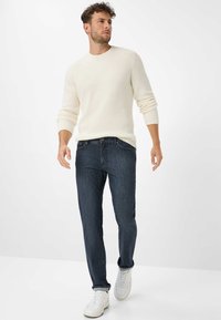 Maglione a coste di colore crema con maniche lunghe abbinato a jeans di denim blu scuro e sneakers bianche. Il modello sta camminando in un'ambientazione neutra.