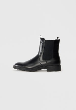 Botas Chelsea de cuero negro con paneles laterales elásticos, puntera redondeada y tacón bajo en bloque. Presentan lengüetas para ponerlas y soltarlas fácilmente.