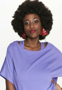 Luźna fioletowa koszulka z krótkim rękawem, nałożona na niebieską bluzkę na ramiączkach. Modelka ma duże czerwone kwiatowe kolczyki i teksturowaną, obfitą fryzurę.