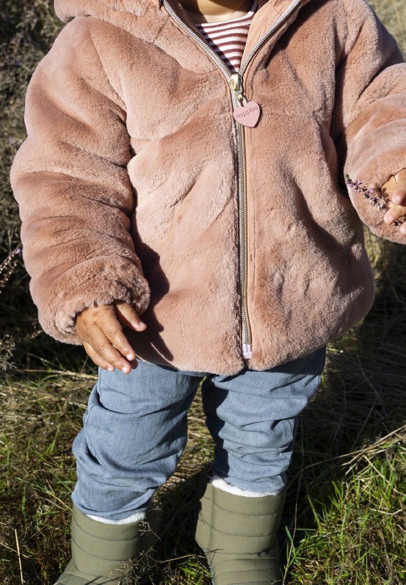 Enfant portant une veste rose duveteuse, un T-shirt rayé, un pantalon bleu et des bottes vertes, debout dans l'herbe, tenant une petite fleur violette.