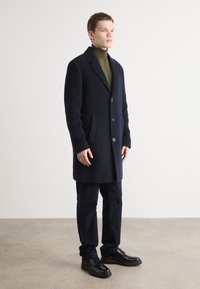 Navyblauer Wollmantel mit taillierter Passform, einreihiger Knopfverschluss und Seitentaschen. Kombiniert mit dunklen Hosen und schwarzen Schuhen.