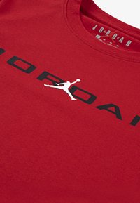 Punainen puuvillapaita, jossa on musta "JORDAN" -teksti ja valkoinen Jumpman-logo. Pyöreä kaula-aukko ja lyhyet hihat, pehmeä pinta.