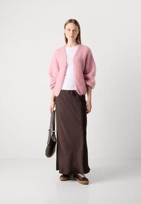 Cardigan rosa fluffy, top bianco aderente, gonna maxi in raso marrone, sneakers marroni con lacci crema e borsa a spalla marrone.