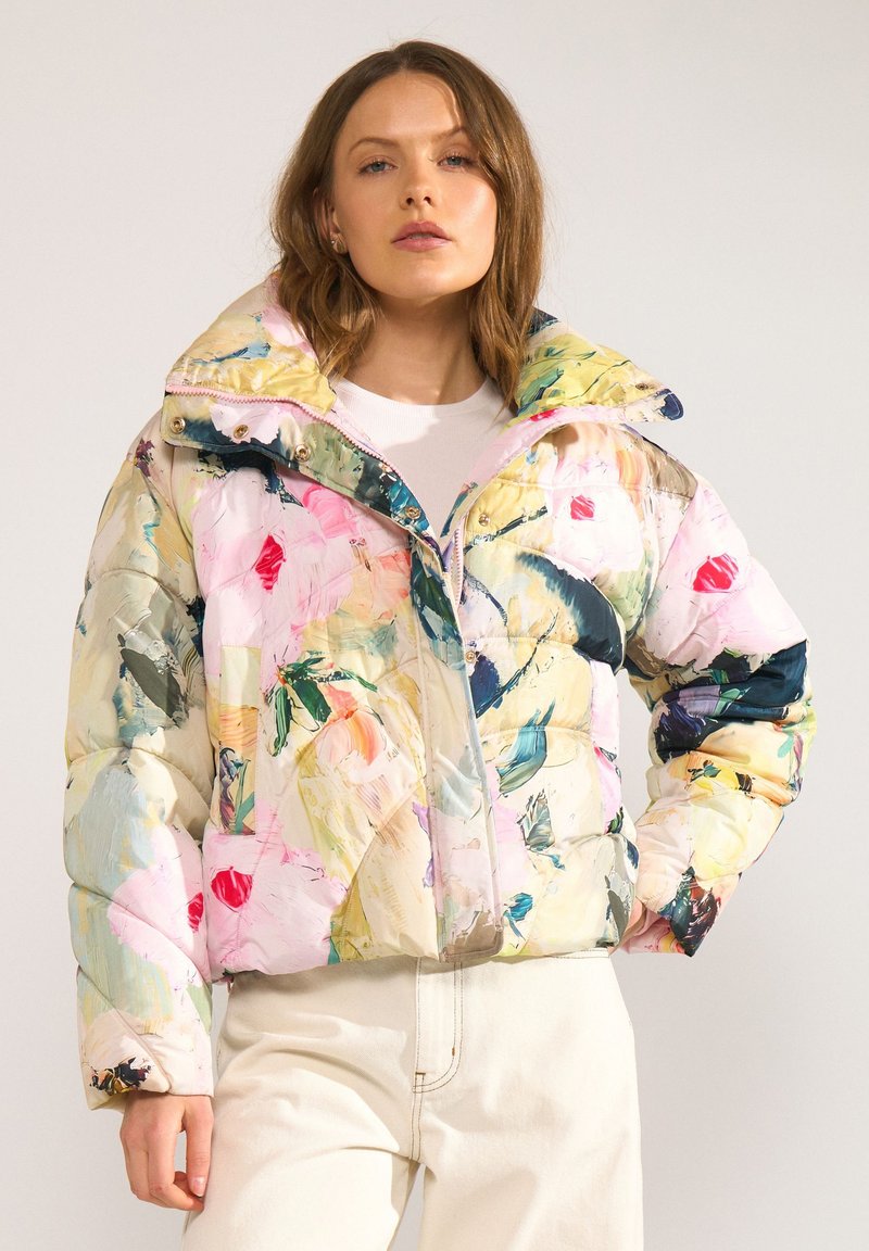 Femme portant une doudoune colorée à motifs floraux avec un col montant et un pantalon clair sur un fond uni.