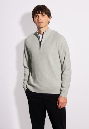 Maglione grigio chiaro con zip, caratterizzato da una texture a costine, con colletto alto e zip nera, abbinato a pantaloni scURI.