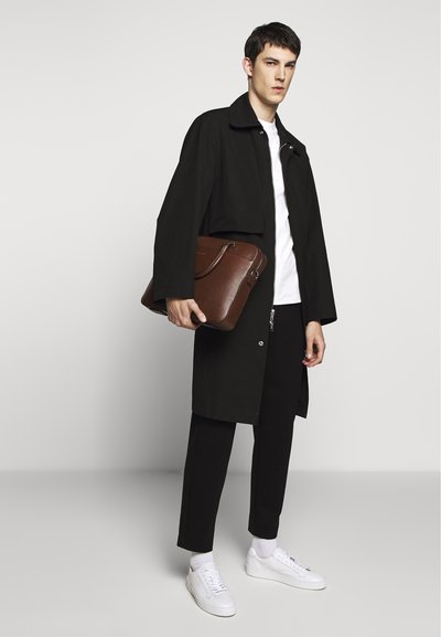 Manteau noir jusqu'à la taille, t-shirt blanc, pantalon noir et baskets blanches. Tenant un sac en cuir texturé marron avec une courte poignée.