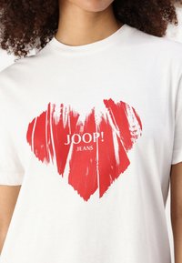 Balta medvilninė marškinėlių palaidinė, kurią puošia didelis raudonas šepetėliu nupieštas širdies piešinys, o centre užrašyta "JOOP! JEANS". Trumpomis rankovėmis, apvaliu iškirpimu.