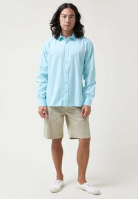 Camisa de manga larga de color azul claro con botones en el frente y cuello, combinada con pantalones cortos tipo cargo beige. El modelo lleva zapatillas blancas; el tejido parece suave.