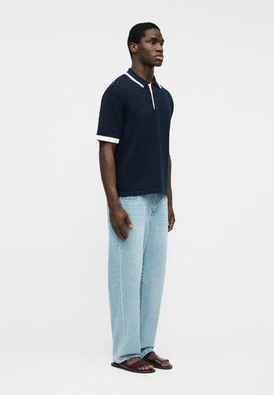 Άνδρας που φοράει μπλε ναυτικό πόλο με λευκή περίγραμμα, ανοιχτό μπλε τζιν και σκούρα καφέ σανδάλια, στέκεται σε λευκό φόντο.