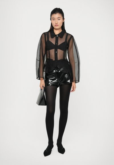 Čierna priesvitná košeľa na gombíky s dlhými rukávmi, v kombinácii s lesklými čiernymi šortkami, čiernymi pančuchami a čiernymi špicatými topánkami, s čiernou taškou v ruke.