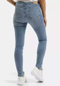 Chude jeansy w jasnoniebieskim denienie, z dwiema tylnymi kieszeniami, przeszytymi szwami oraz widoczną metką marki na pasku. Noszone z białymi trampkami.