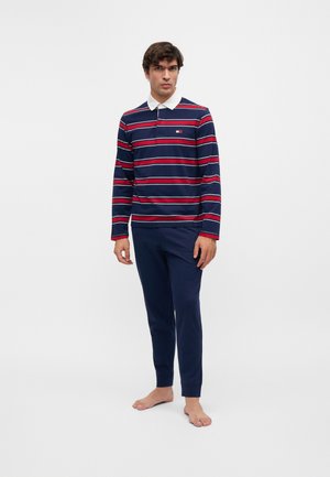 Man die een navyblauw en rood gestreept poloshirt met lange mouwen en een witte kraag draagt en effen navyblauwe broek, staat blootsvoets op een witte achtergrond.