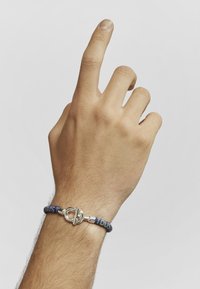 Pulsera de tela en tonos azul marino con un cierre de palanca de plata, que presenta intrincados patrones en azul y amarillo, llevada en una mano levantada.