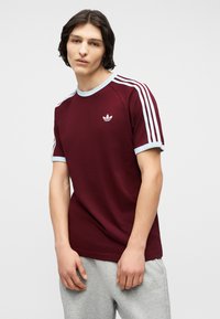 Νεαρός άνδρας με σκούρα μαλλιά μέχρι τον ώμο, ντυμένος με μπορντό μπλουζάκι Adidas με γαλάζια λεπτομέρεια και γκρι παντελόνι φόρμας, μπροστά σε απλό φόντο.